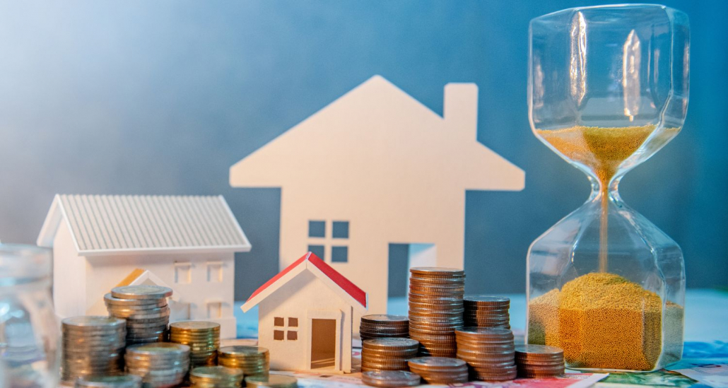 Hausmodell mit Sanduhr und Münzen als Symbol für Wert und Zeitpunkt (Immobilienbewertung Hohenbrunn)